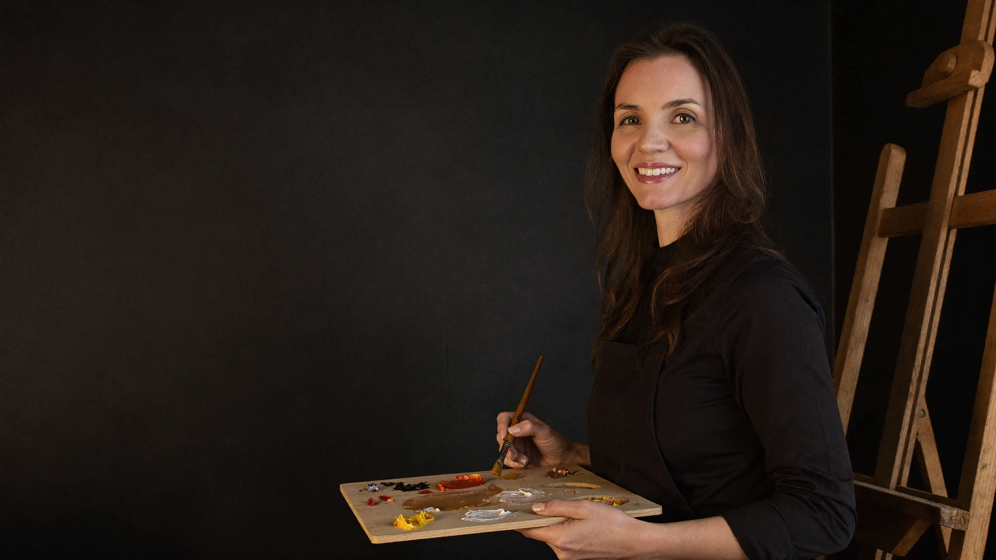 Lari Namaste em seu ateliê, com paleta de tintas acrílicas e pincel na mão, ao lado do cavalete