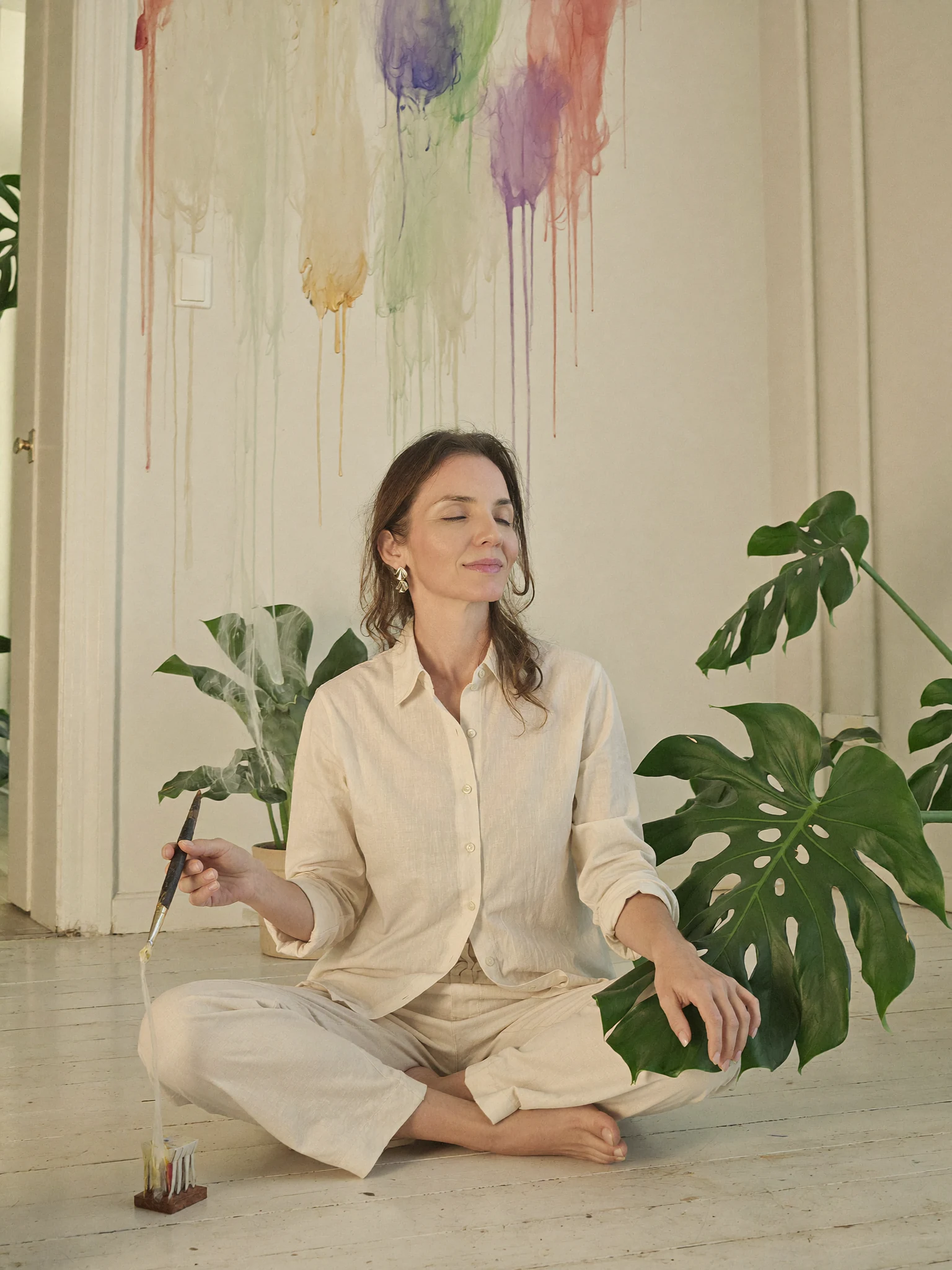 Lari Namaste sentada no chão do ateliê, serena, ao lado de uma folha de costela-de-adão, com tinta colorida escorrendo na parede